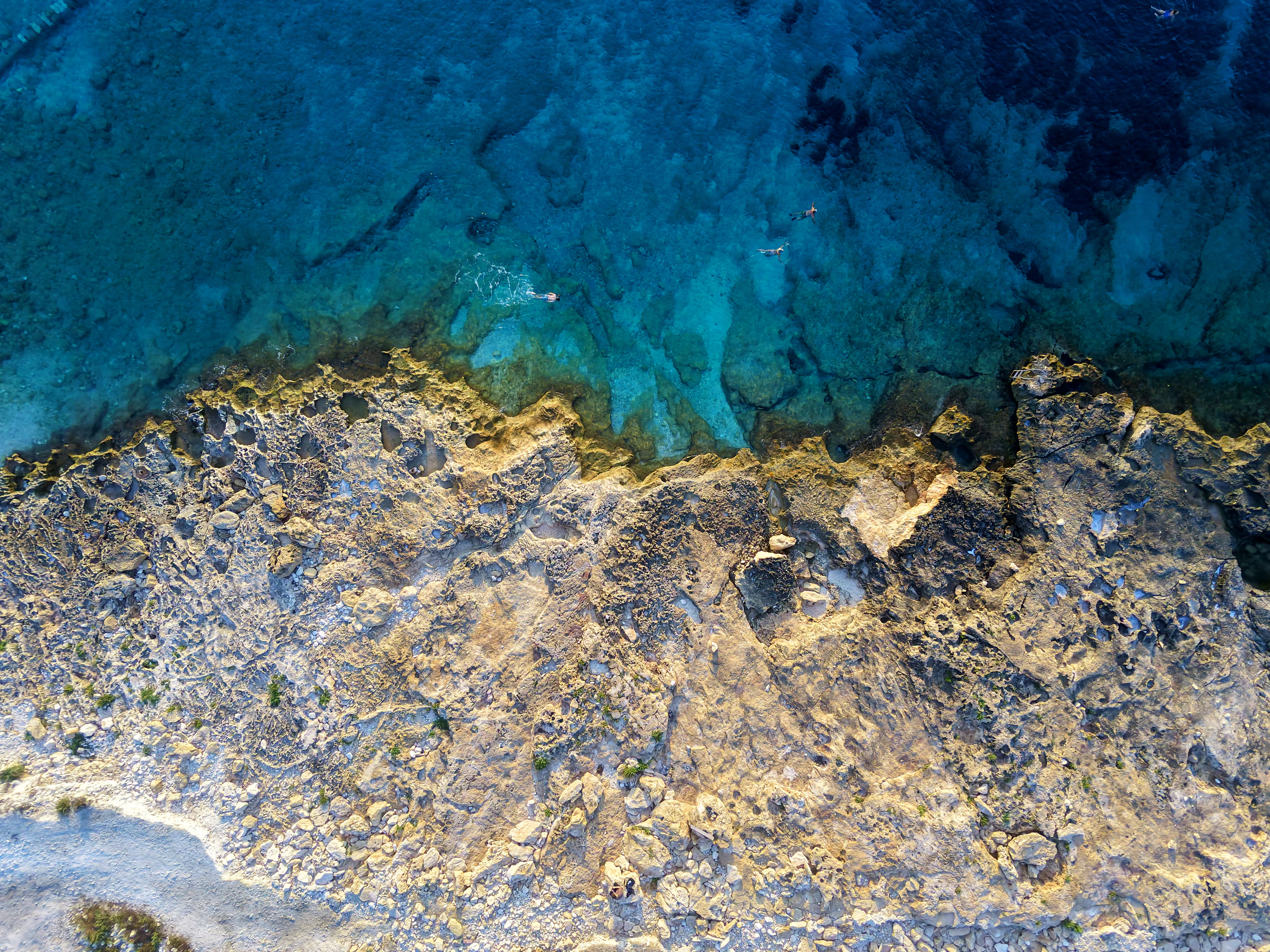 Swimmers over turquoise shallows where the rocky shore meets the sea at Ħondoq ir-Rummien, Gozo, seen from directly above
